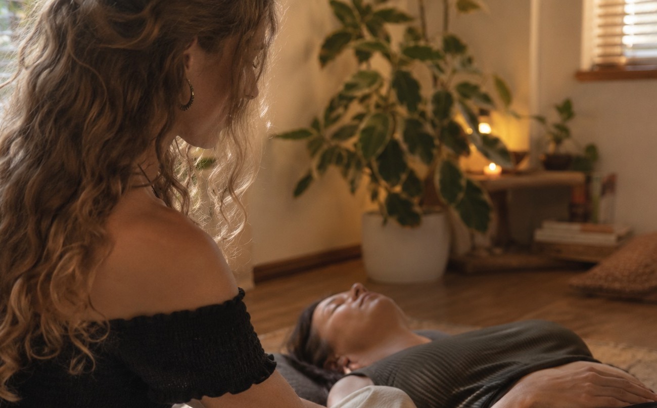 Anna-Lena sitting beside a client lying down, resting her hands gently on the client's arm in a warm, softly lit room
